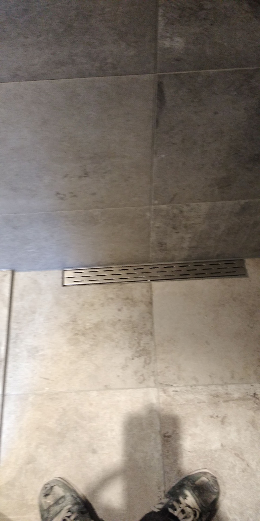 Szary odpływ liniowy wkomponowany w jasne płytki podłogowe w łazience, widoczne stopy w butach w dolnej części kadru.