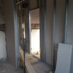 Montaż metalowego stelaża pod płyty gipsowo-kartonowe w surowym wnętrzu, widoczny laser krzyżowy na statywie, drewniane kantówki i fragment ściany z cegły.