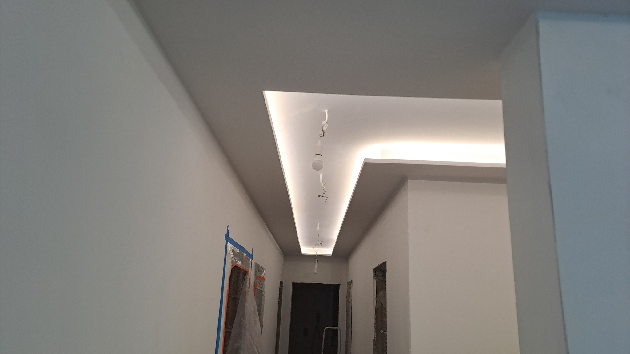 Korytarz w trakcie remontu z podwieszonym sufitem z oświetleniem LED, ściany przygotowane do malowania, zabezpieczone drzwi folią malarską.