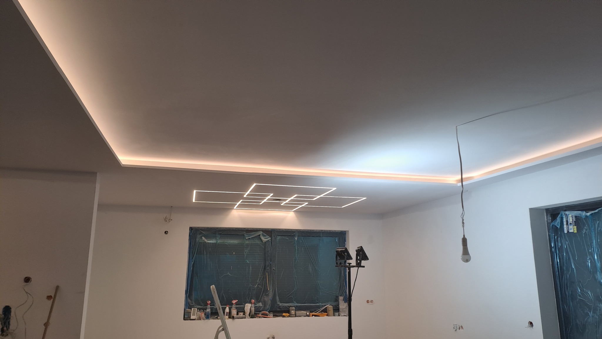 Nowoczesne oświetlenie sufitowe z listwami LED i geometrycznym wzorem z profili aluminiowych. Okno zasłonięte folią ochronną, widoczne ślady prac wykończeniowych i drabina.