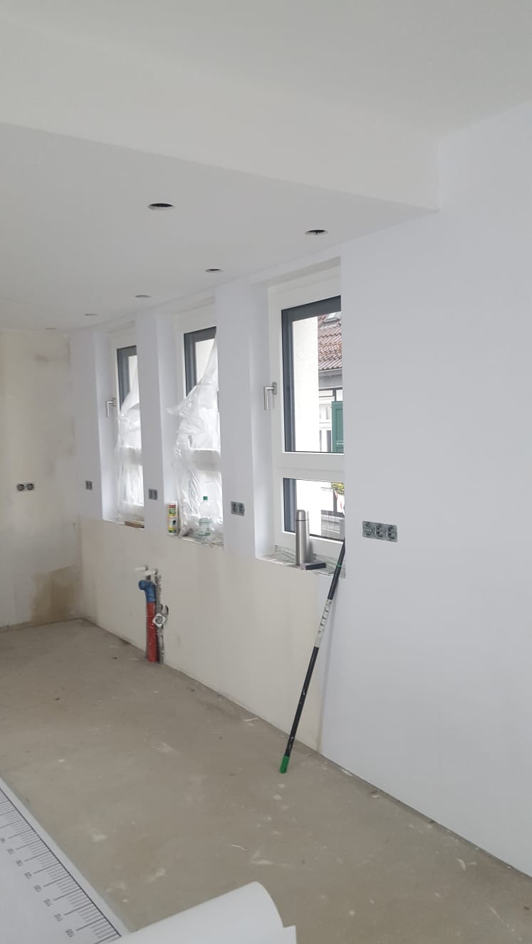 Wnętrze w trakcie prac wykończeniowych: świeżo pomalowane na biało ściany, zakryte folią okna, widoczne puszki instalacyjne i wałek do malowania oparty o ścianę.