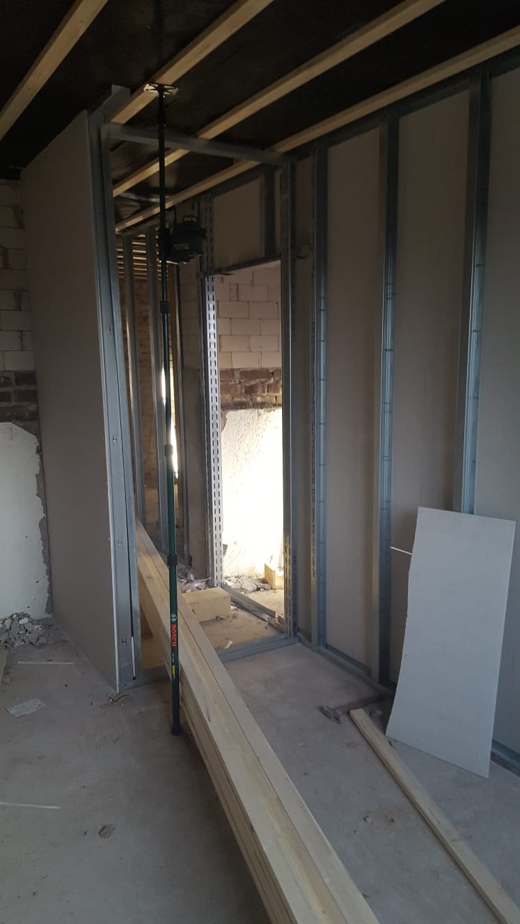 Montaż metalowego stelaża pod płyty gipsowo-kartonowe w surowym wnętrzu, widoczny laser krzyżowy na statywie, drewniane kantówki i fragment ściany z cegły.