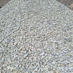 Stos szarego kruszywa o nieregularnych kształtach, z bliska, na tle zieleni drzew i fragmentu murowanego ogrodzenia.