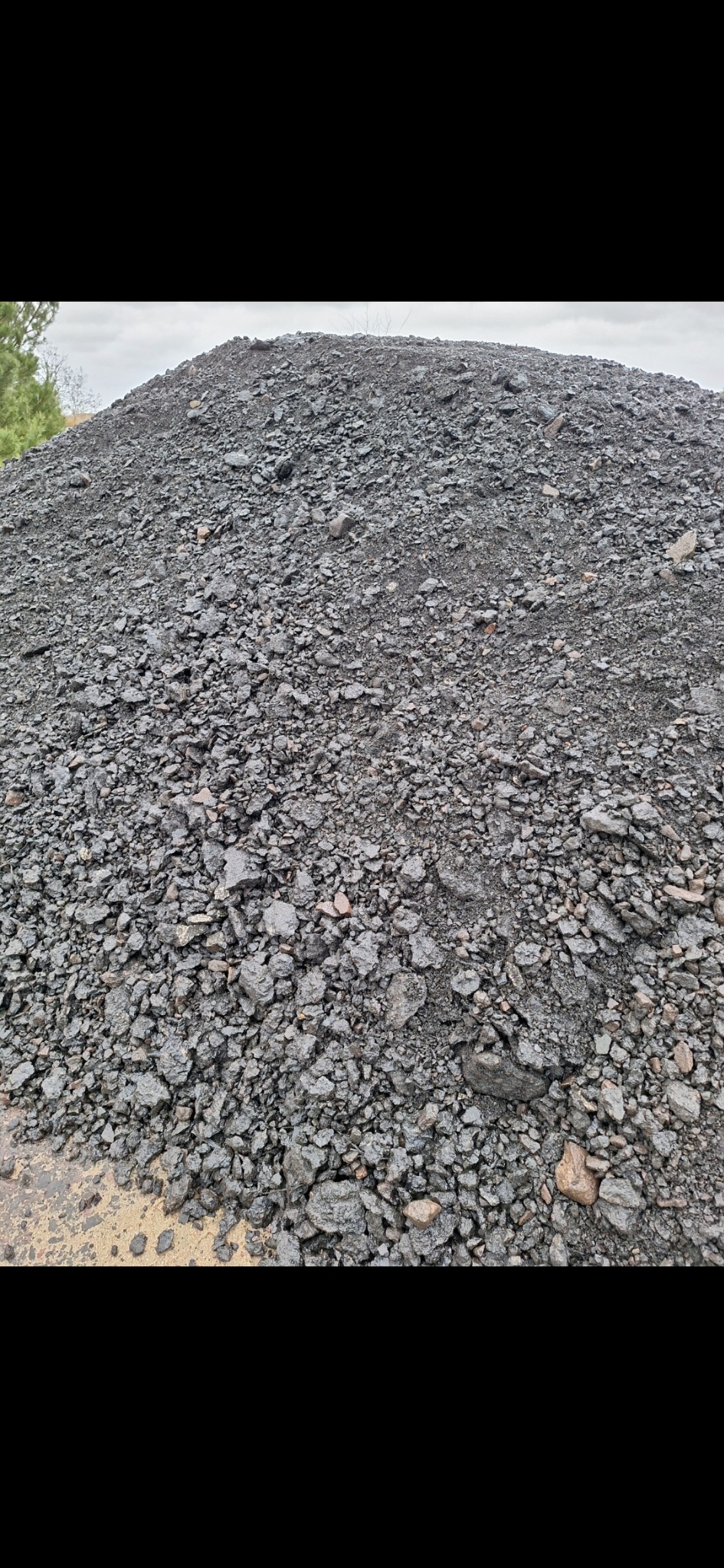 Góra wilgotnego, ciemnoszarego kruszywa na placu budowy, widoczne pojedyncze brązowe kamienie i częściowo żółte podłoże.