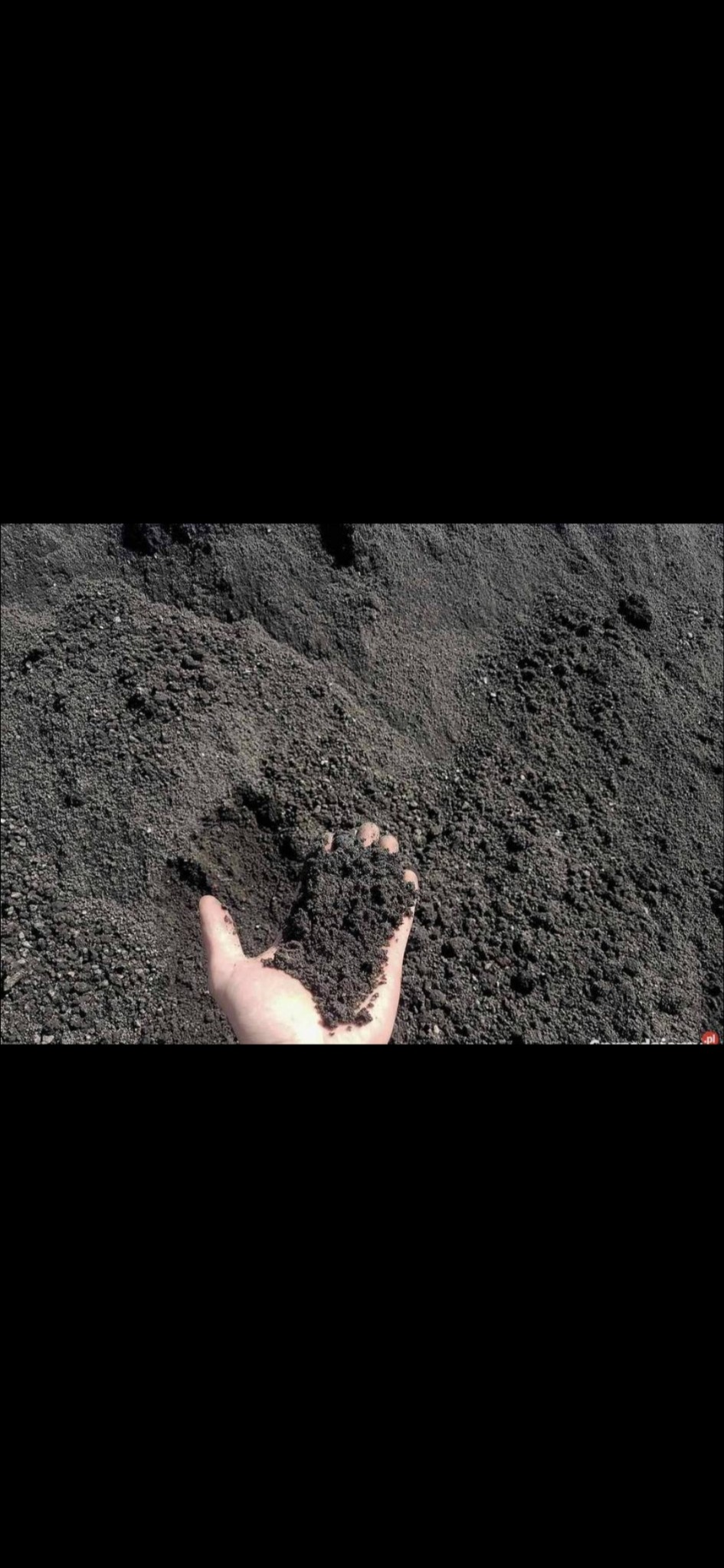 Dłoń trzymająca ciemny piasek na tle pryzmy tego samego materiału, widoczne drobne grudki i nierówności powierzchni.