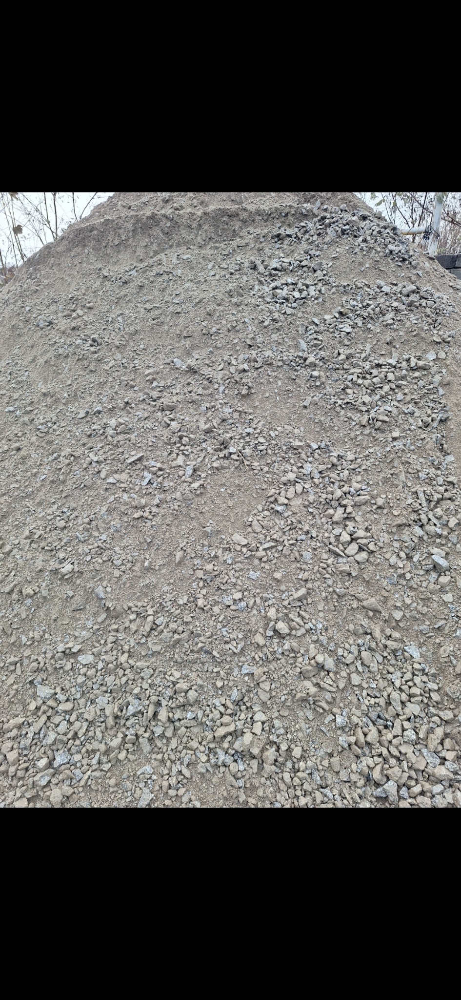 Kupa szarego kruszywa o zróżnicowanej granulacji, widoczna struktura piasku i drobnych kamieni, na zewnątrz, w tle drzewa bez liści.