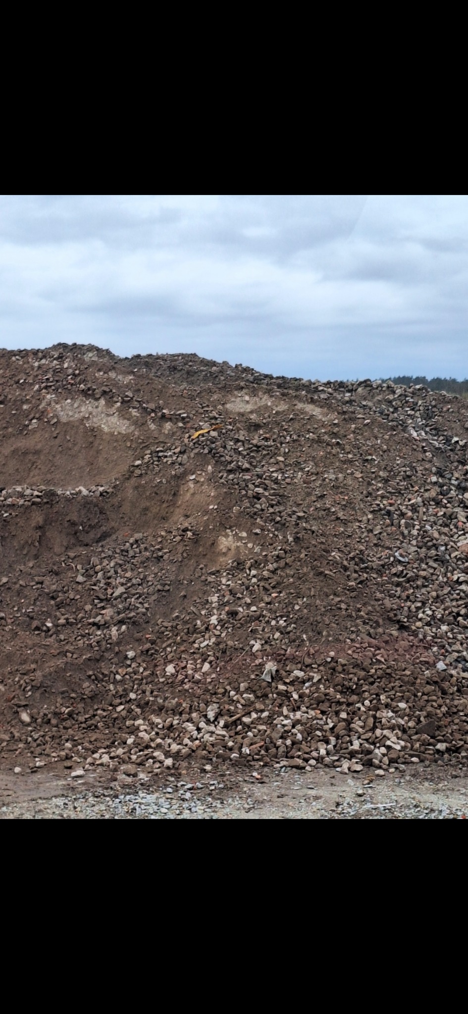 Góra gruzu i ziemi pod pochmurnym niebem, widoczny fragment żółtej taśmy ostrzegawczej na szczycie.