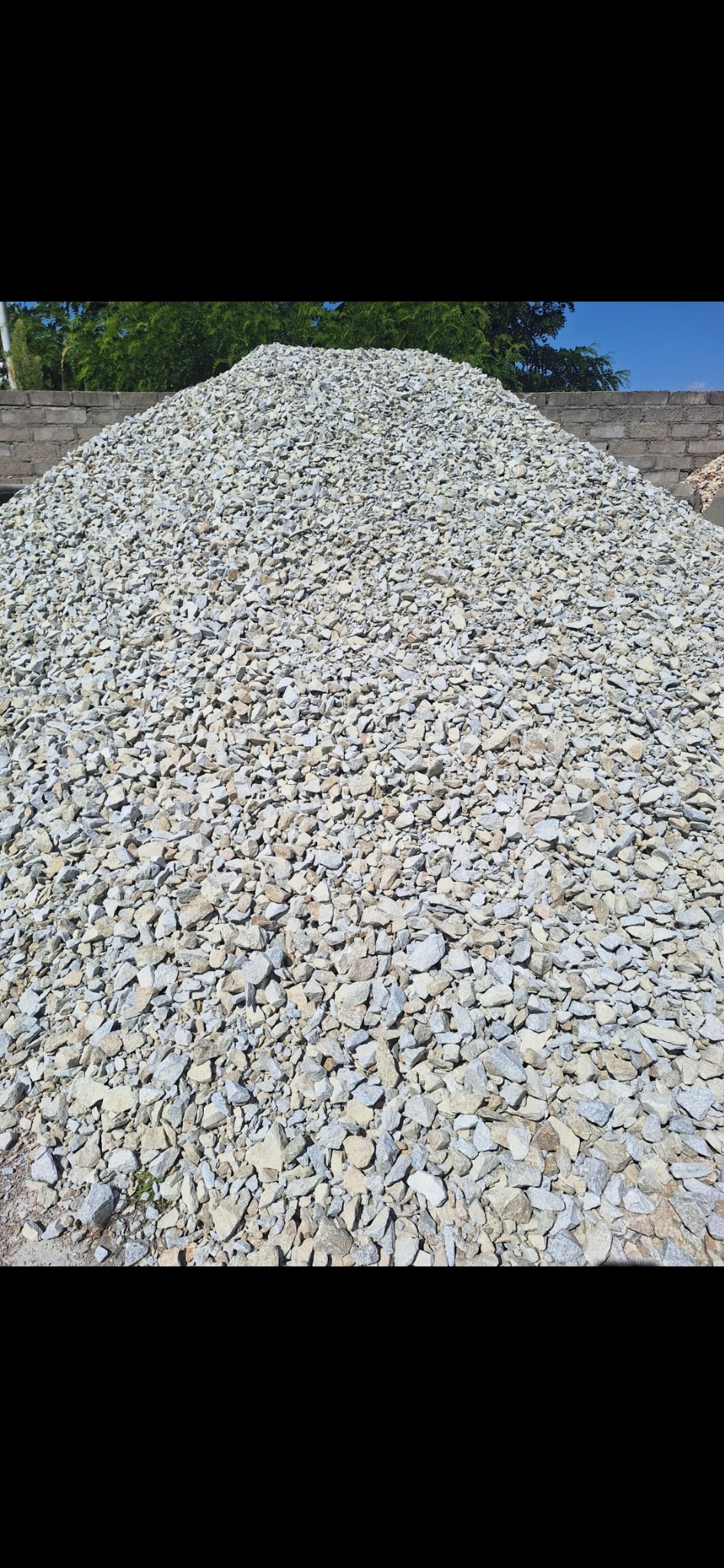 Stos szarego kruszywa o nieregularnych kształtach, z bliska, na tle zieleni drzew i fragmentu murowanego ogrodzenia.