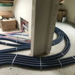 Wnętrze w trakcie instalacji systemu wentylacyjnego: rury wentylacyjne w czarnych karbowanych osłonach, przymocowane metalowymi taśmami, prowadzone po surowej podłodze, widoczny rozdzielacz...