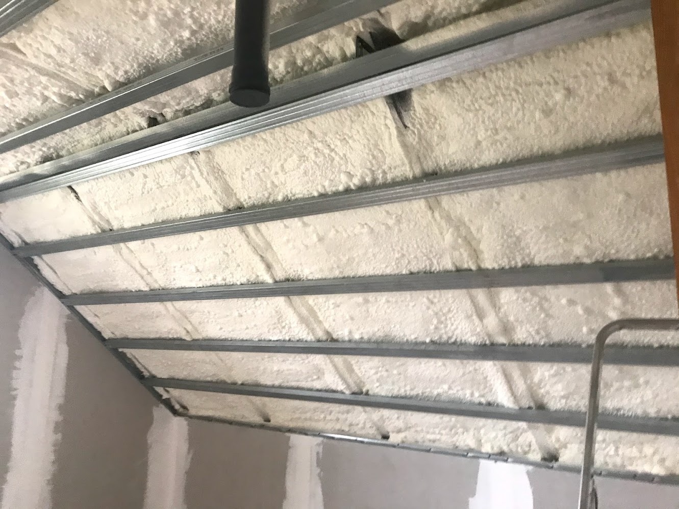 Poddasze ocieplone białą pianką natryskową, widoczne metalowe profile konstrukcyjne sufitu i fragment rury. Ściana boczna częściowo wykończona gładzią.