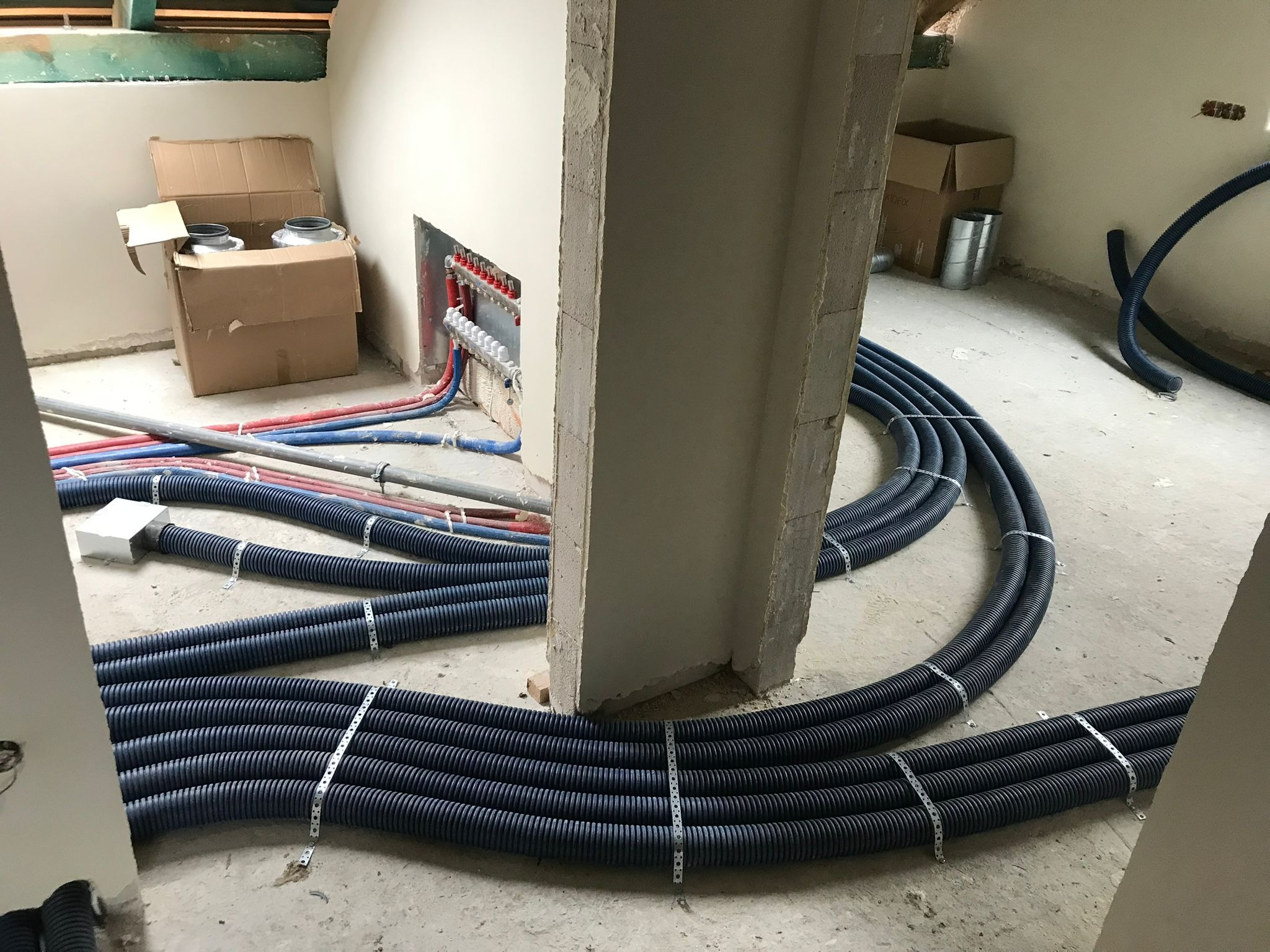 Wnętrze w trakcie instalacji systemu wentylacyjnego: rury wentylacyjne w czarnych karbowanych osłonach, przymocowane metalowymi taśmami, prowadzone po surowej podłodze, widoczny rozdzielacz...