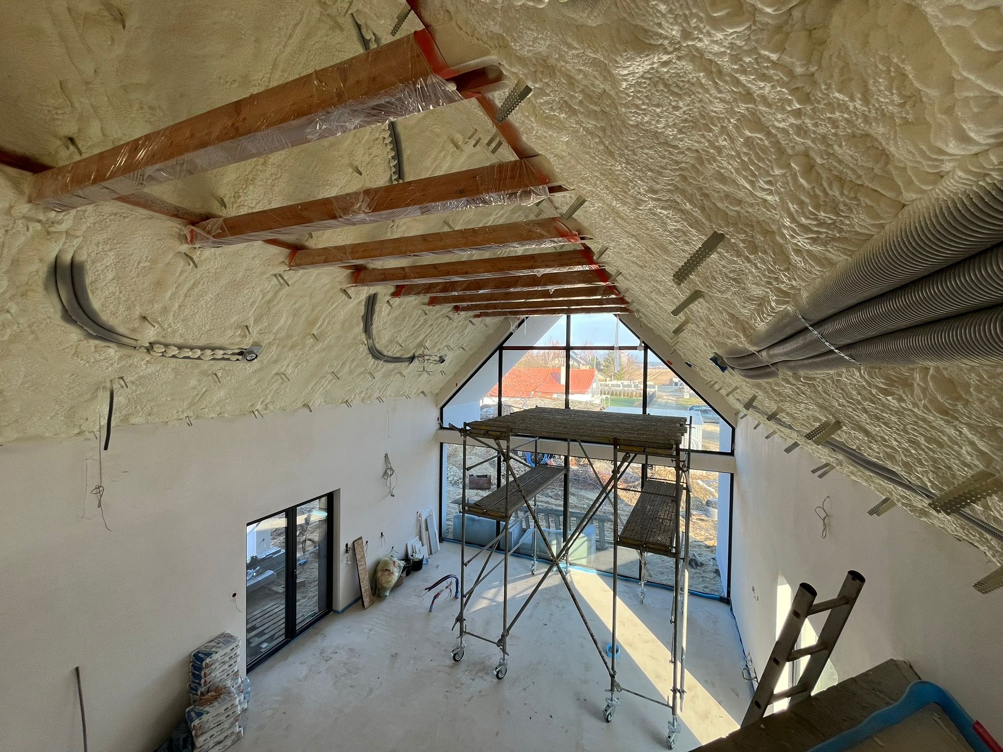 Poddasze domu jednorodzinnego po ociepleniu pianką PUR, widoczne drewniane elementy konstrukcyjne zabezpieczone folią, rusztowanie i worki z materiałem budowlanym na podłodze, duże okno z widokiem...