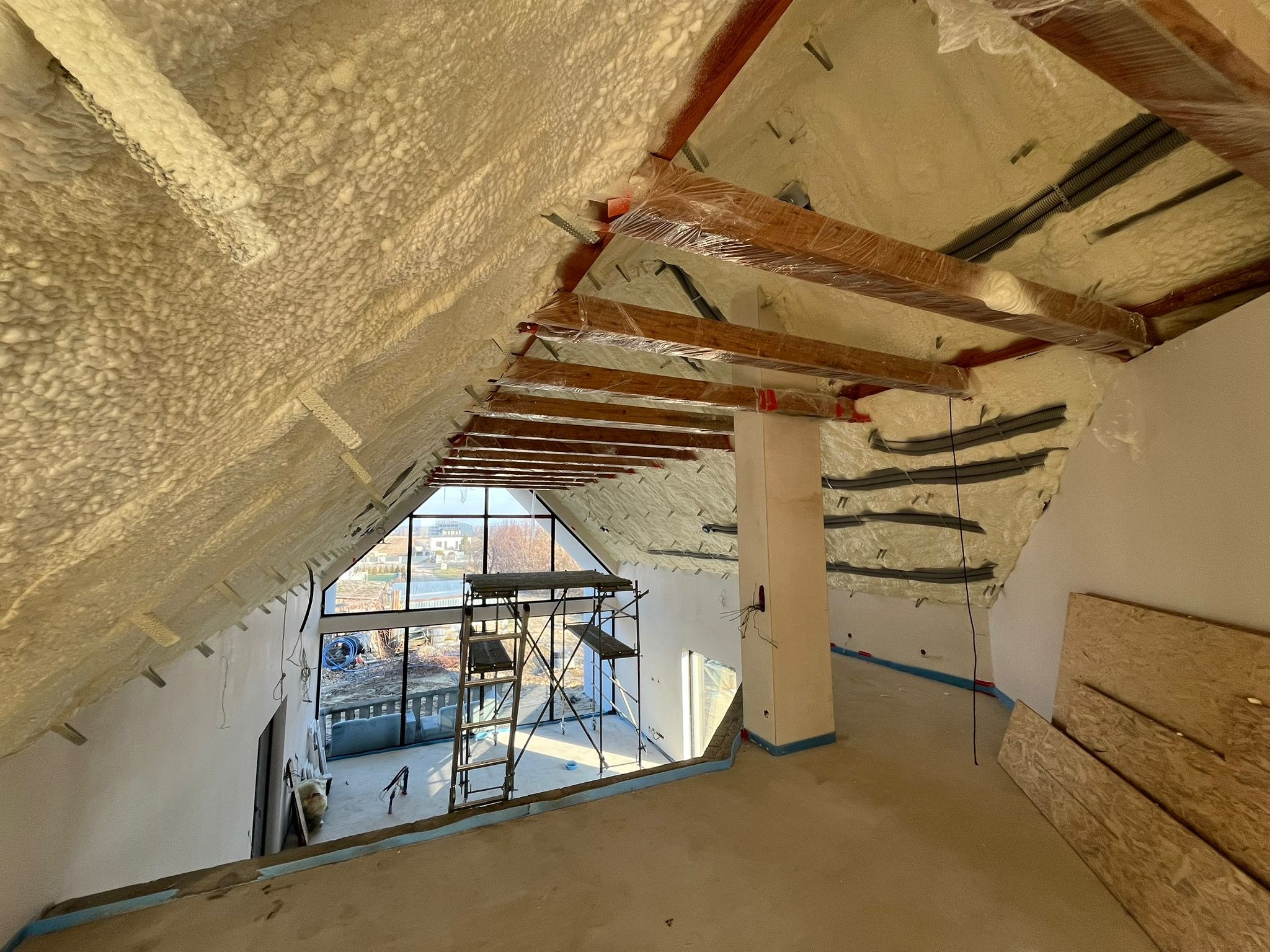 Poddasze domu jednorodzinnego w trakcie prac izolacyjnych, widoczna warstwa pianki izolacyjnej na skośnych ścianach i suficie, drewniane belki zabezpieczone folią, rusztowanie przy oknie...