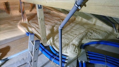 Poddasze z widoczną izolacją natryskową, drewnianą konstrukcją więźby dachowej i instalacją elektryczną w niebieskich peszlach.