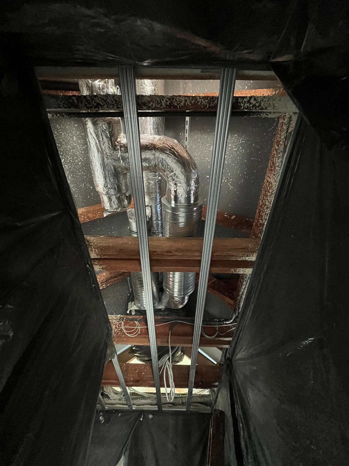 Widok z dołu na system wentylacyjny z rurami w izolacji aluminiowej, zamontowany pod drewnianym stropem, z widocznymi elementami konstrukcyjnymi i przewodami elektrycznymi, częściowo zasłonięty...