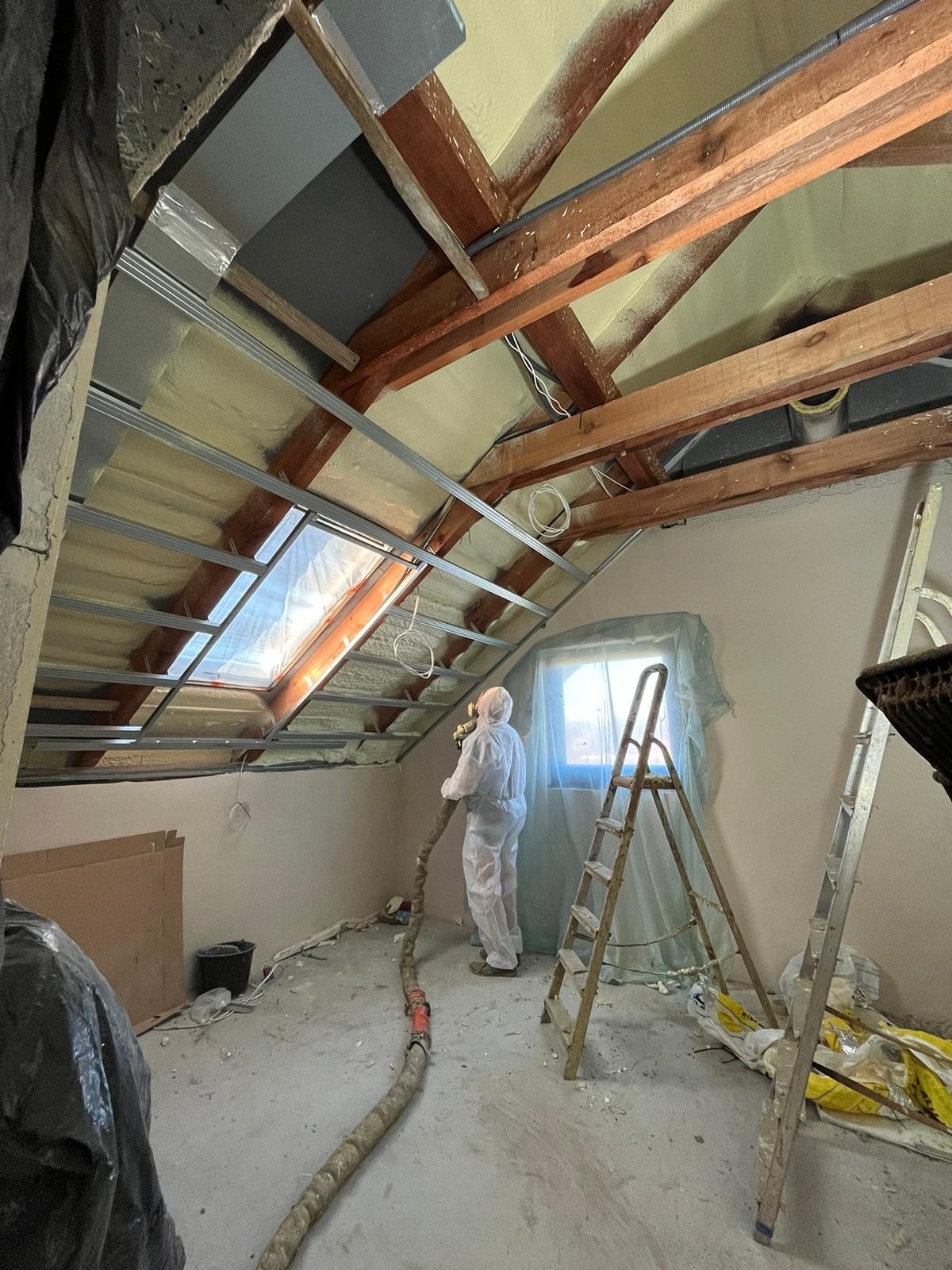 Pracownik w kombinezonie ochronnym aplikuje izolację natryskową na poddaszu z oknem dachowym, widoczne drewniane belki, metalowe profile, kable elektryczne, oraz sprzęt do natrysku izolacji.