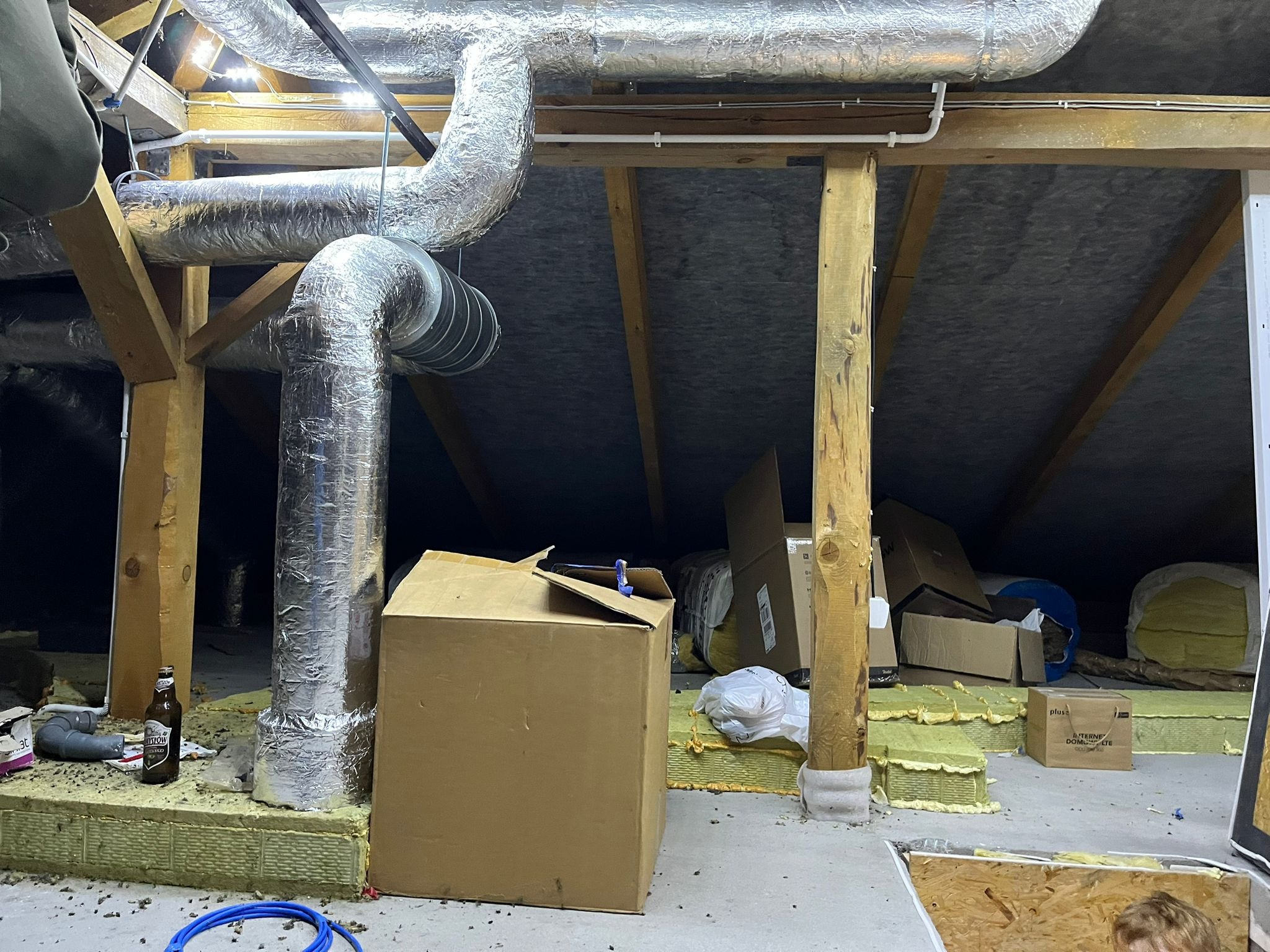 Poddasze z widocznymi elementami instalacji wentylacyjnej z rurami w srebrnej izolacji, częściowo ocieplone wełną mineralną, z kartonami i butelką piwa. Widoczne drewniane elementy konstrukcyjne...