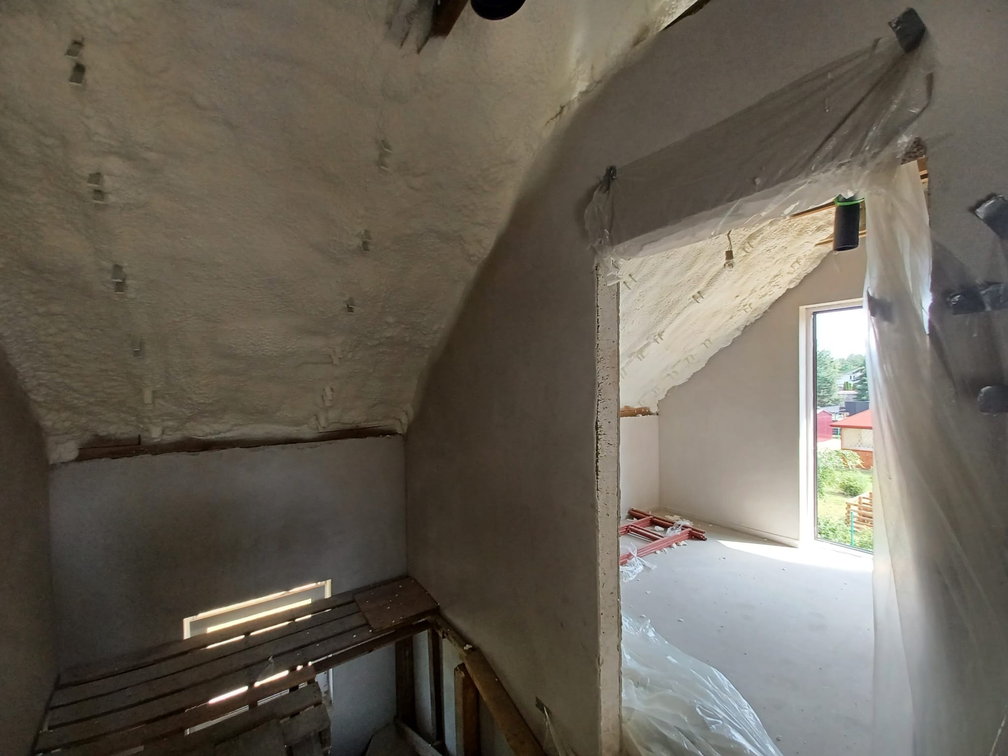 Poddasze w trakcie ocieplania pianką PUR, widoczne metalowe mocowania w pianie, fragment podestu roboczego i okno z widokiem na zieleń.