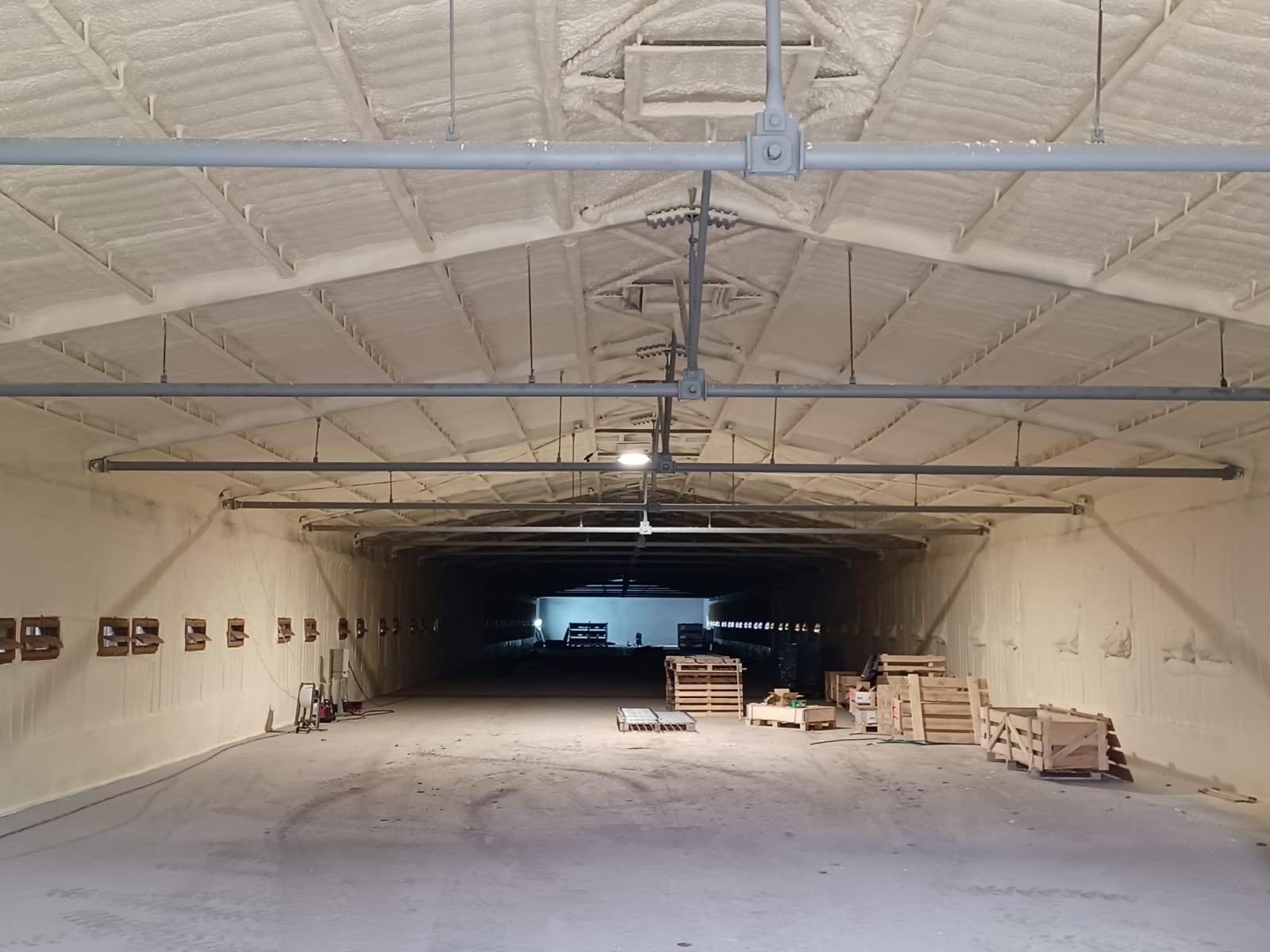 Wnętrze hali przemysłowej po ociepleniu pianką, widoczne szare rury pod sufitem i drewniane palety na podłodze, ściany i sufit pokryte grubą warstwą pianki izolacyjnej.