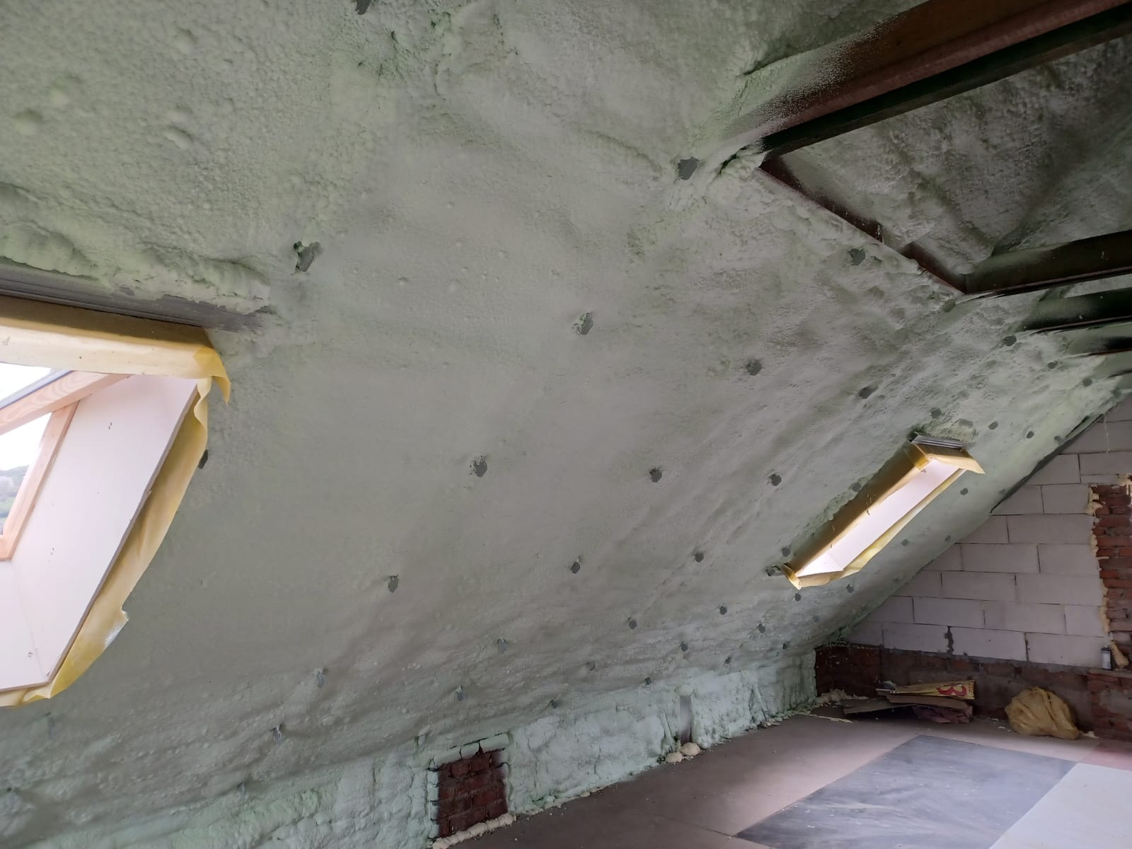 Poddasze po ociepleniu pianką PUR, widoczne okna dachowe zabezpieczone taśmą malarską, ślady po kołkach montażowych w piance, fragment ściany z pustaków i cegły w dolnej części kadru.