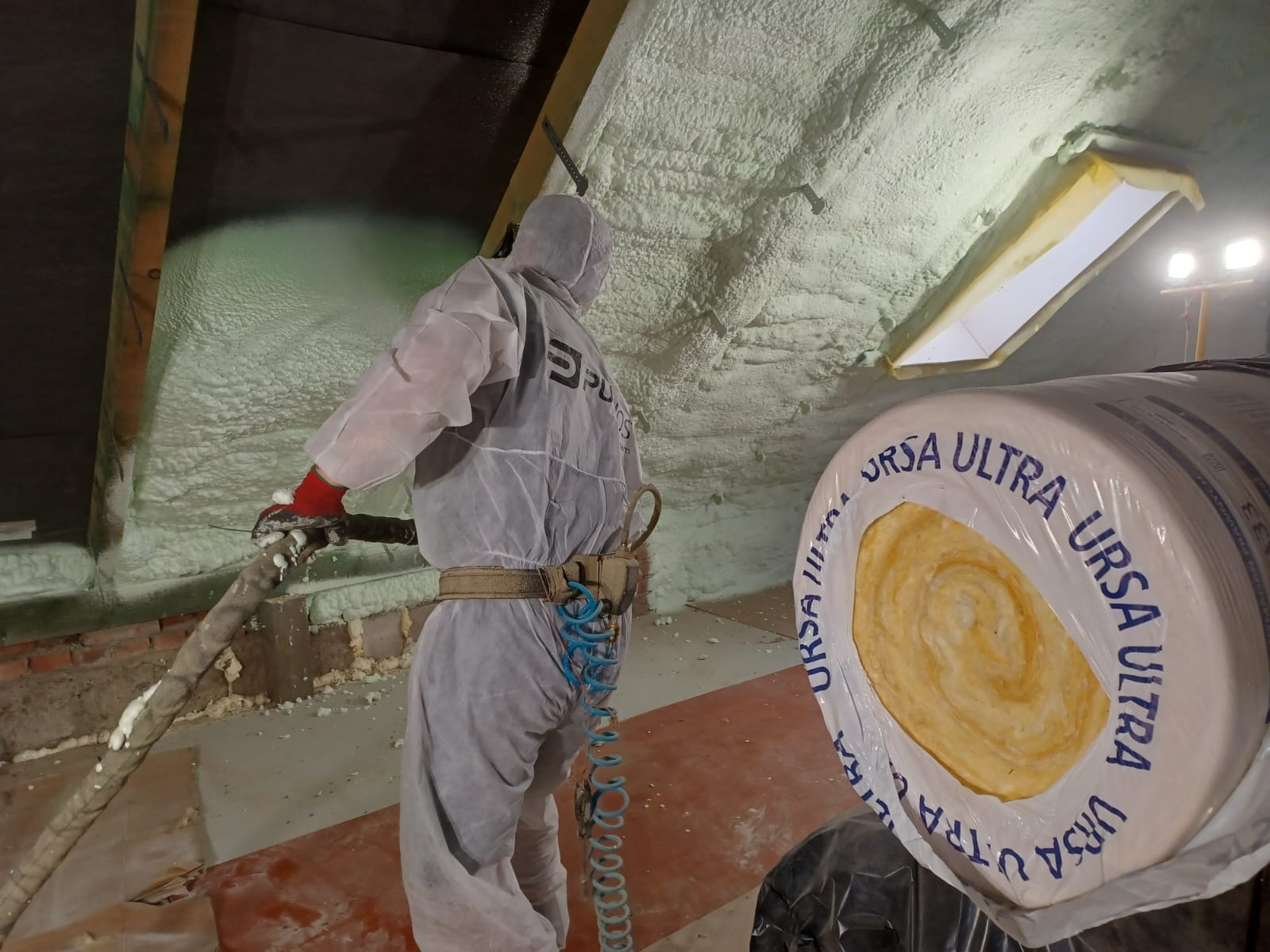 Pracownik w kombinezonie ochronnym aplikuje piankę izolacyjną na poddaszu, widoczny fragment okna dachowego i rolka wełny mineralnej Ursa Ultra.