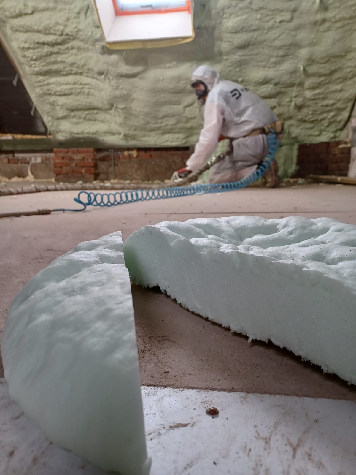 Pracownik w kombinezonie ochronnym aplikuje pianę na poddaszu, widoczny fragment dociętej piany w pierwszym planie, niebieski wąż od pistoletu natryskowego.