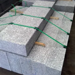 Szare krawężniki granitowe ułożone w stosy, zabezpieczone zielonymi taśmami transportowymi i drewnianymi przekładkami.