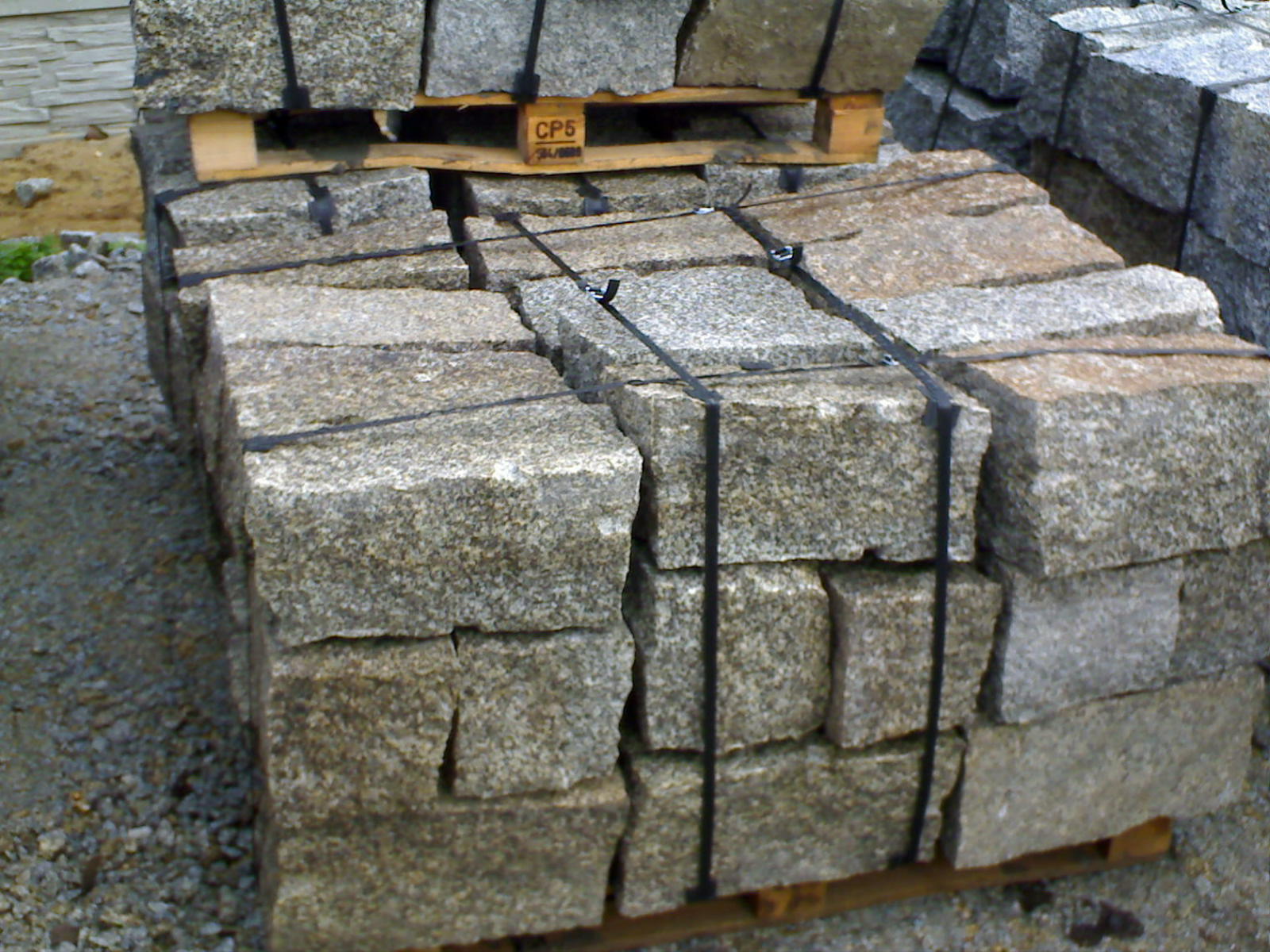 Palety z kamieniem murowym, zabezpieczone czarną taśmą, ułożone na żwirowym podłożu.