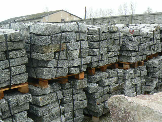 Szare, granitowe kostki murowe ułożone w stosy na drewnianych paletach, zabezpieczone czarnymi taśmami, z fragmentem większego kamienia w prawym dolnym rogu i betonowym ogrodzeniem w tle.