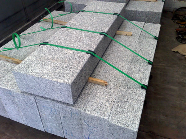 Szare krawężniki granitowe ułożone w stosy, zabezpieczone zielonymi taśmami transportowymi i drewnianymi przekładkami.