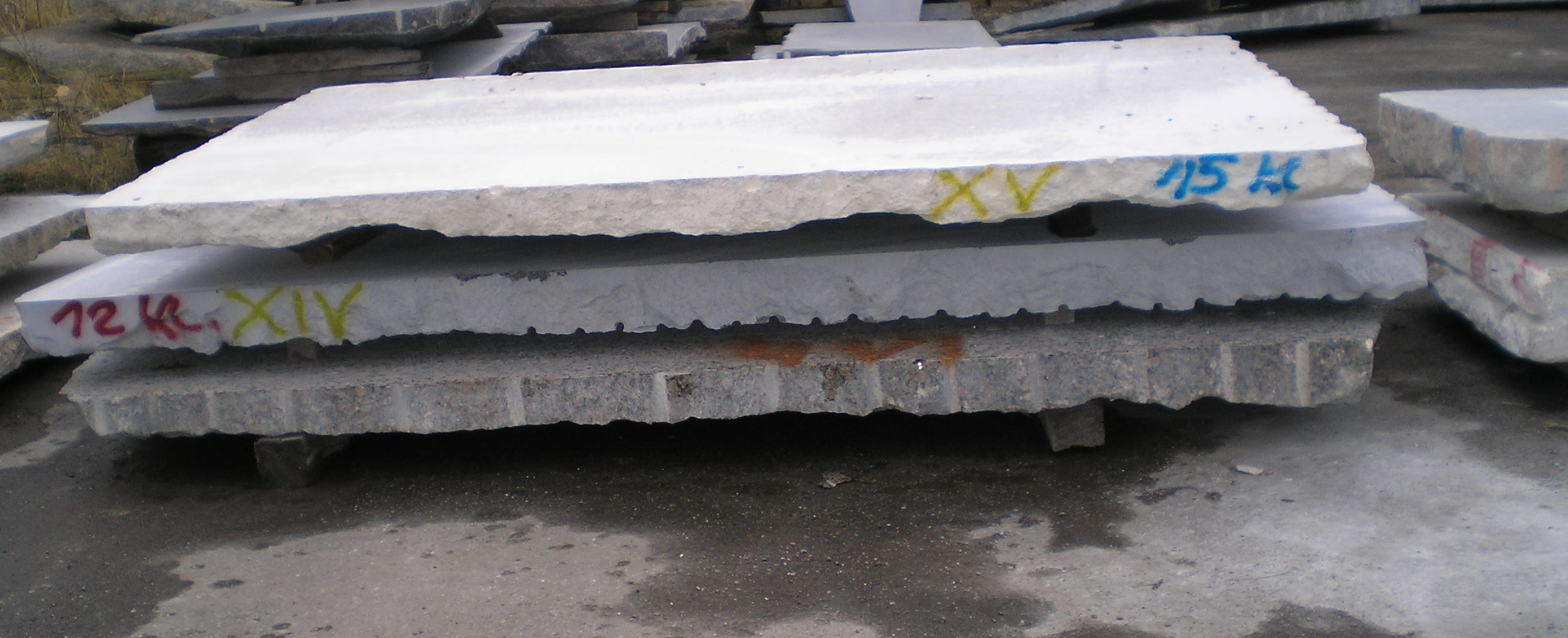 Stos szarych płyt granitowych o nieregularnych krawędziach, z widocznymi oznaczeniami numeracji w kolorze żółtym, czerwonym i niebieskim, leżących na betonowej powierzchni.