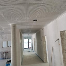 KW PRO-BUD - Wnętrze w trakcie prac wykończeniowych: sufit podwieszany z perforacją, kolumna w surowym stanie, ściany przygotowane do malowania, widoczny sprzęt i materiały budowlane.