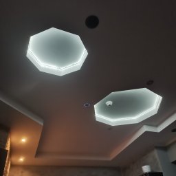 KW PRO-BUD - Sufit z dwoma ośmiokątnymi wnękami podświetlonymi taśmą LED, dodatkowe oświetlenie punktowe i czujnik dymu, widoczne fragmenty umeblowania i dekoracji wnętrza.