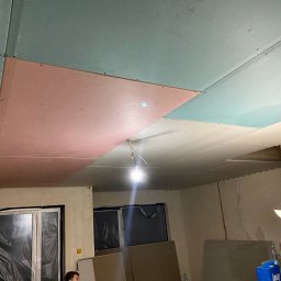 KW PRO-BUD - Montaż sufitu podwieszanego z płyt kartonowo-gipsowych w pomieszczeniu, widoczne płyty w różnych kolorach (różowy, zielony, biały) przed szpachlowaniem i malowaniem, wisząca lampa budowlana.