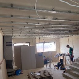 KW PRO-BUD - Montaż sufitu podwieszanego z płyt kartonowo-gipsowych, widoczne profile metalowe, płyty gipsowe opierające się o ścianę, paleta z narzędziami na podłodze, pracownik w tle.