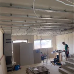 Montaż sufitu podwieszanego z płyt kartonowo-gipsowych, widoczne profile metalowe, płyty gipsowe opierające się o ścianę, paleta z narzędziami na podłodze, pracownik w tle.