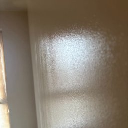 KW PRO-BUD - Świeżo pomalowana, beżowa ściana z widoczną fakturą farby i odbijającym się światłem, fragment okna zabezpieczonego folią malarską w lewym dolnym rogu.