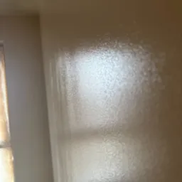 Świeżo pomalowana, beżowa ściana z widoczną fakturą farby i odbijającym się światłem, fragment okna zabezpieczonego folią malarską w lewym dolnym rogu.