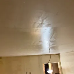 Świeżo nałożona, mokra warstwa gładzi na suficie z widocznymi pęknięciami i niedoskonałościami, surowa ściana w tle z widocznymi śladami po taśmie malarskiej, kabel zwisający z sufitu, oświetlenie...