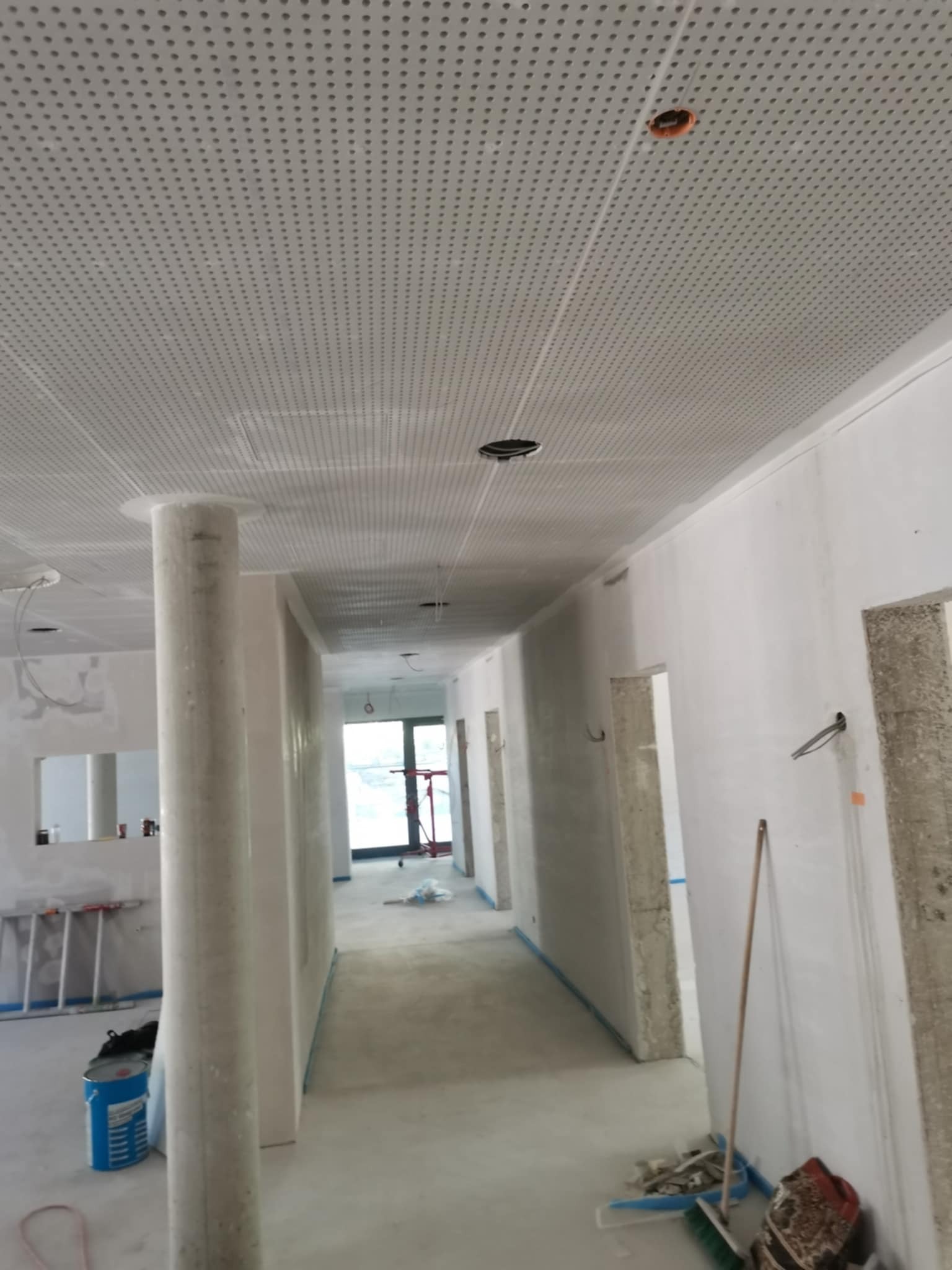 Wnętrze w trakcie prac wykończeniowych: sufit podwieszany z perforacją, kolumna w surowym stanie, ściany przygotowane do malowania, widoczny sprzęt i materiały budowlane.
