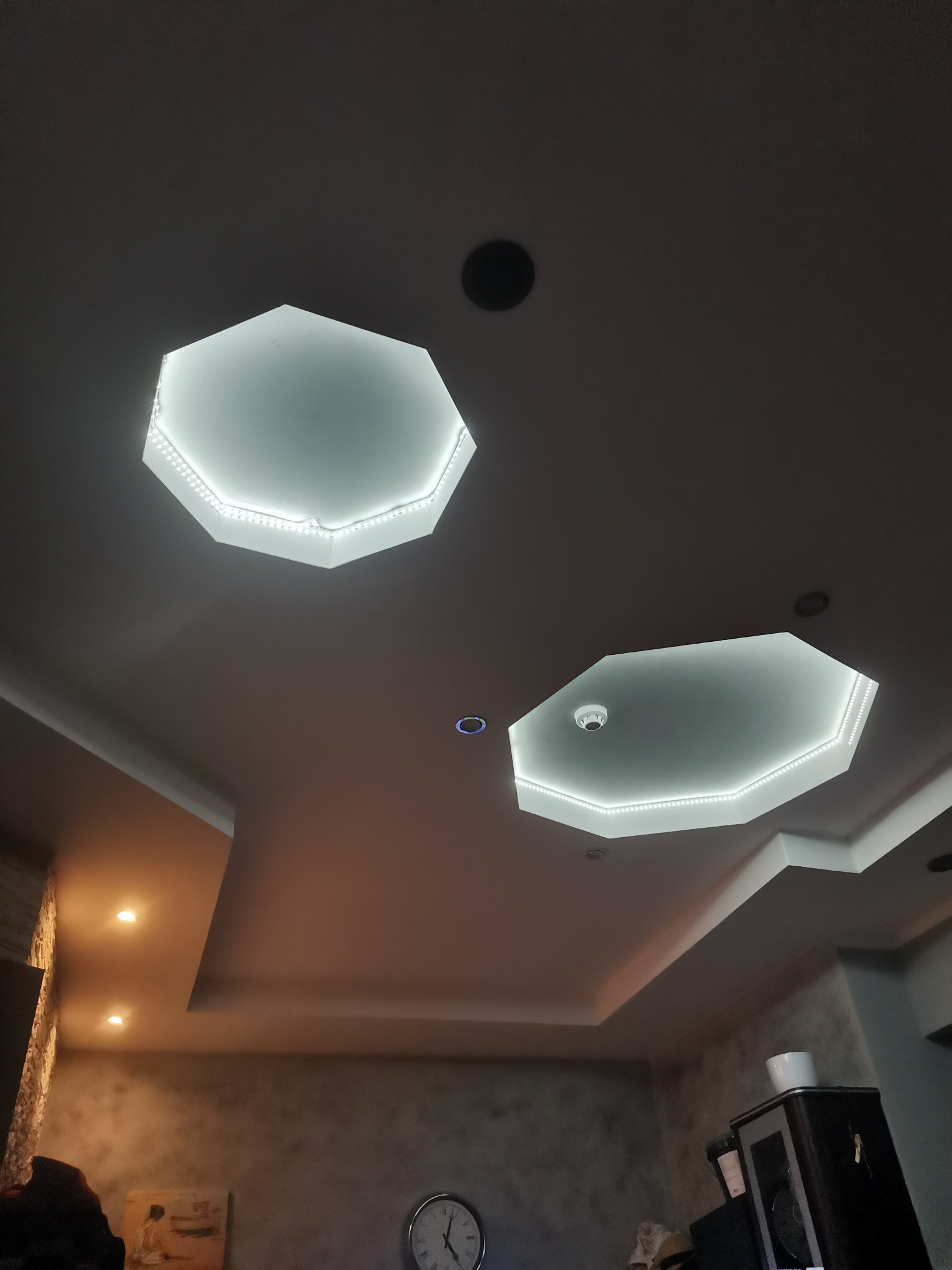 Sufit z dwoma ośmiokątnymi wnękami podświetlonymi taśmą LED, dodatkowe oświetlenie punktowe i czujnik dymu, widoczne fragmenty umeblowania i dekoracji wnętrza.