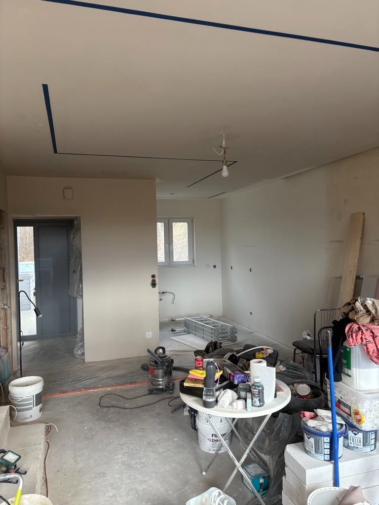 Wnętrze w trakcie prac przygotowawczych: surowy stan ścian, zabezpieczone taśmą malarską krawędzie sufitu, rozstawiona drabina, widoczny stół roboczy z narzędziami i materiałami budowlanymi.