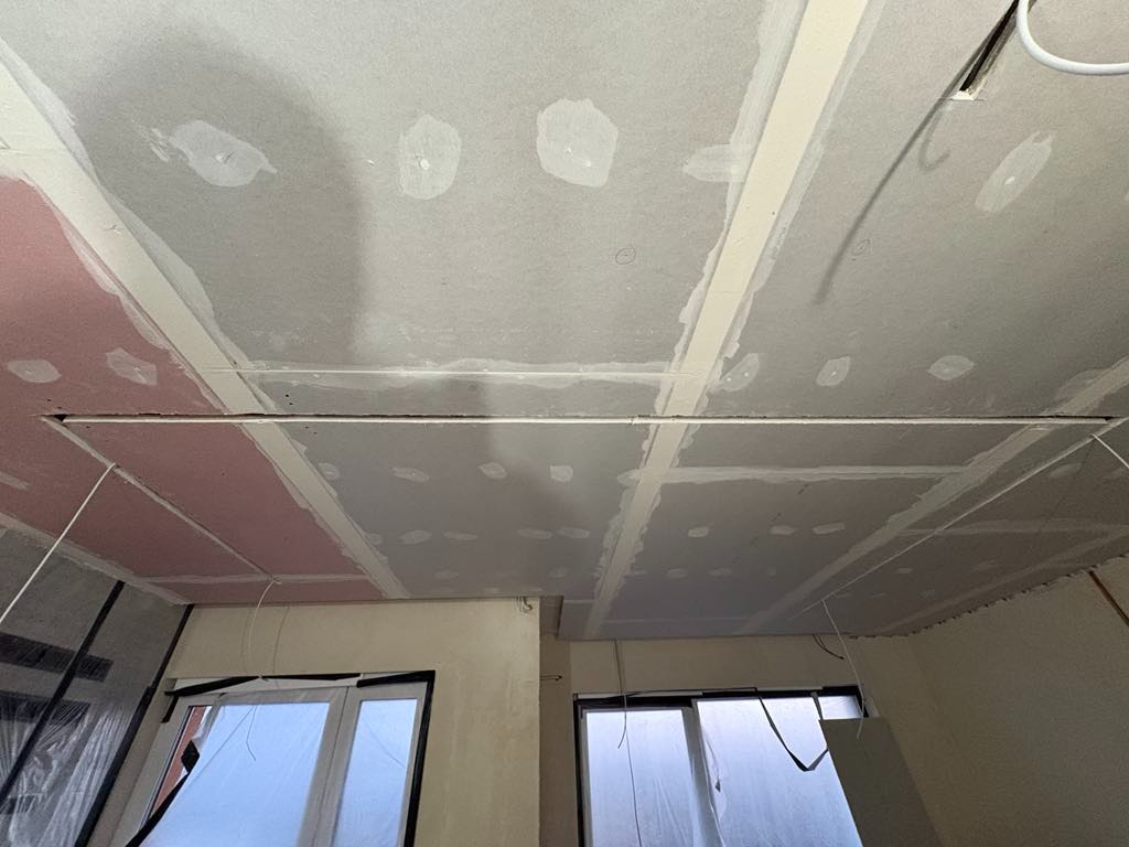 Sufit z płyt kartonowo-gipsowych w trakcie montażu, widoczne łączenia płyt i wstępnie zaszpachlowane miejsca wkrętów, częściowo zabezpieczone okno folią ochronną.