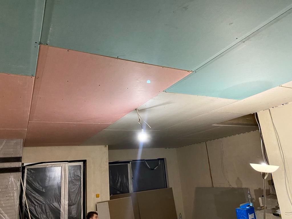 Montaż sufitu podwieszanego z płyt kartonowo-gipsowych w pomieszczeniu, widoczne płyty w różnych kolorach (różowy, zielony, biały) przed szpachlowaniem i malowaniem, wisząca lampa budowlana.