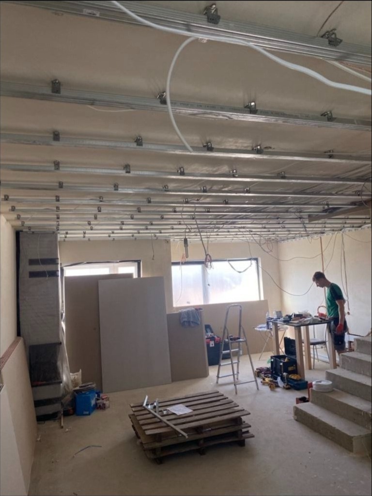 Montaż sufitu podwieszanego z płyt kartonowo-gipsowych, widoczne profile metalowe, płyty gipsowe opierające się o ścianę, paleta z narzędziami na podłodze, pracownik w tle.