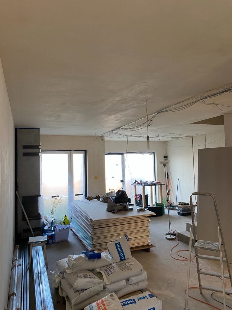 Wnętrze w trakcie remontu: ułożone płyty karton gips, worki z zaprawą, widoczne przewody elektryczne pod sufitem, drabina, szpachlowana ściana i fragment kominka.