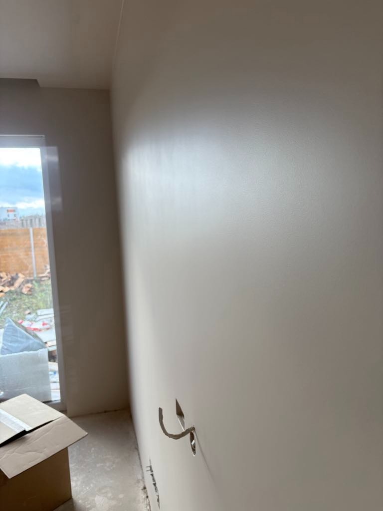 Świeżo pomalowana, gładka ściana w jasnym odcieniu z widocznymi wystającymi przewodami elektrycznymi, karton na podłodze i widokiem przez okno na zewnątrz.