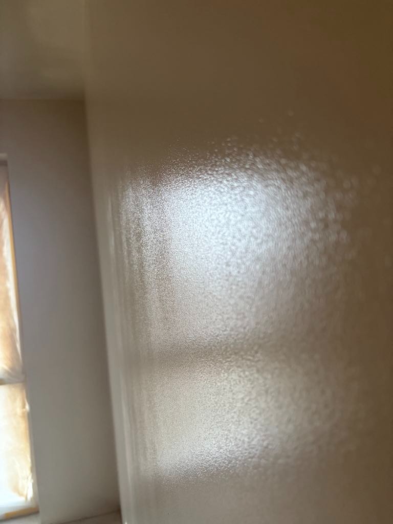 Świeżo pomalowana, beżowa ściana z widoczną fakturą farby i odbijającym się światłem, fragment okna zabezpieczonego folią malarską w lewym dolnym rogu.