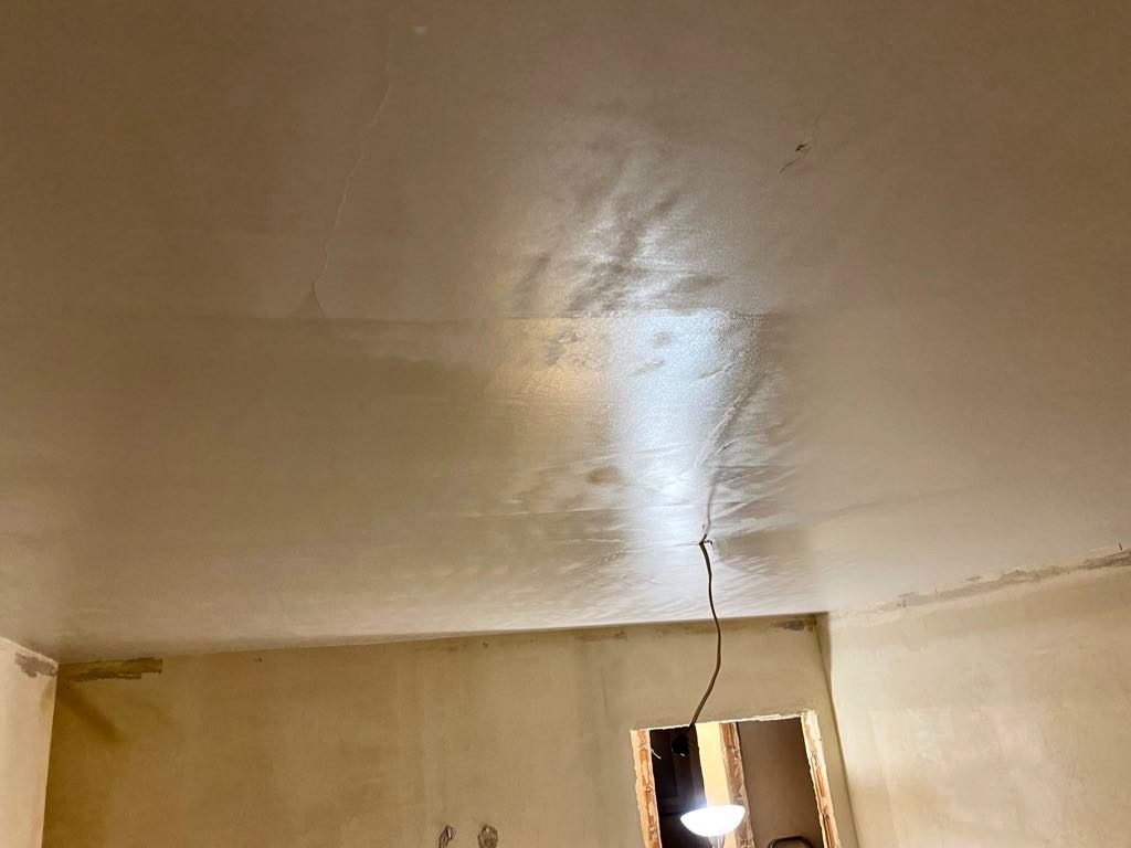 Świeżo nałożona, mokra warstwa gładzi na suficie z widocznymi pęknięciami i niedoskonałościami, surowa ściana w tle z widocznymi śladami po taśmie malarskiej, kabel zwisający z sufitu, oświetlenie...