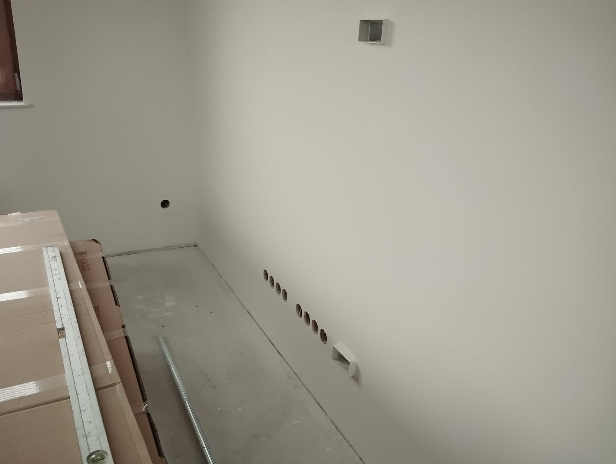 Świeżo wywiercone otwory w ścianie, przygotowane do montażu punktów oświetleniowych, widoczne kartony z elementami wyposażenia i poziomica.